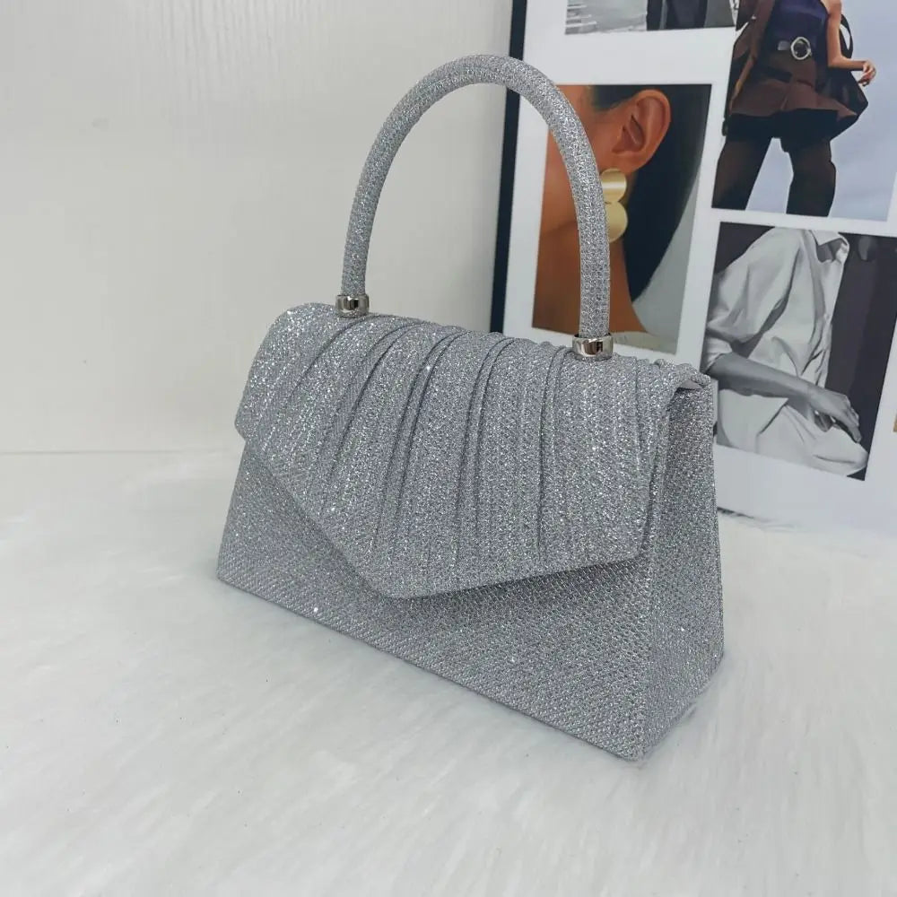 A silver glitter textured handbag with a handle is displayed in front of a photo backdrop.