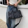A woman wearing a white top and jeans holds a black saddle-shaped PU leather city tote bag against a white wall.
