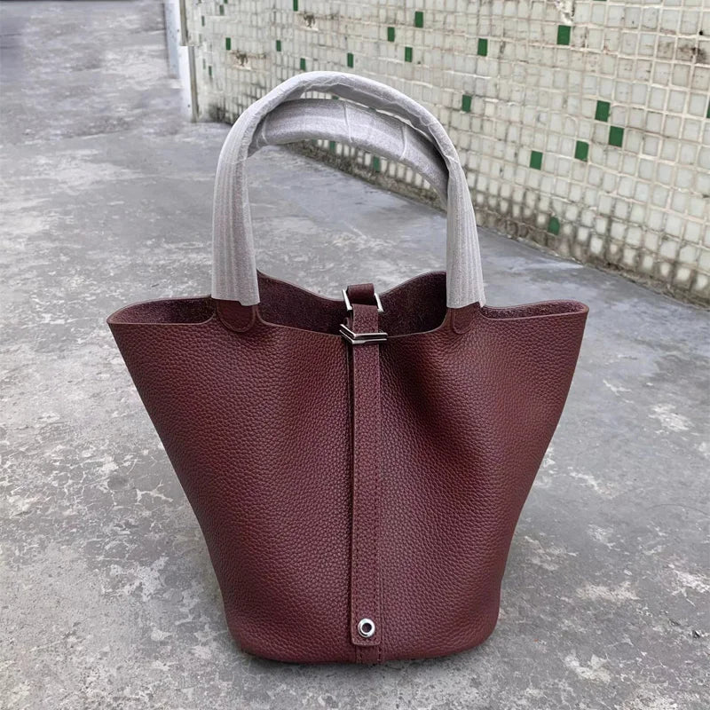 Ein tiefburgunderes Leder-Bucket-Bag mit einem hellgrauen Stoffgriff auf einer Betonoberfläche.