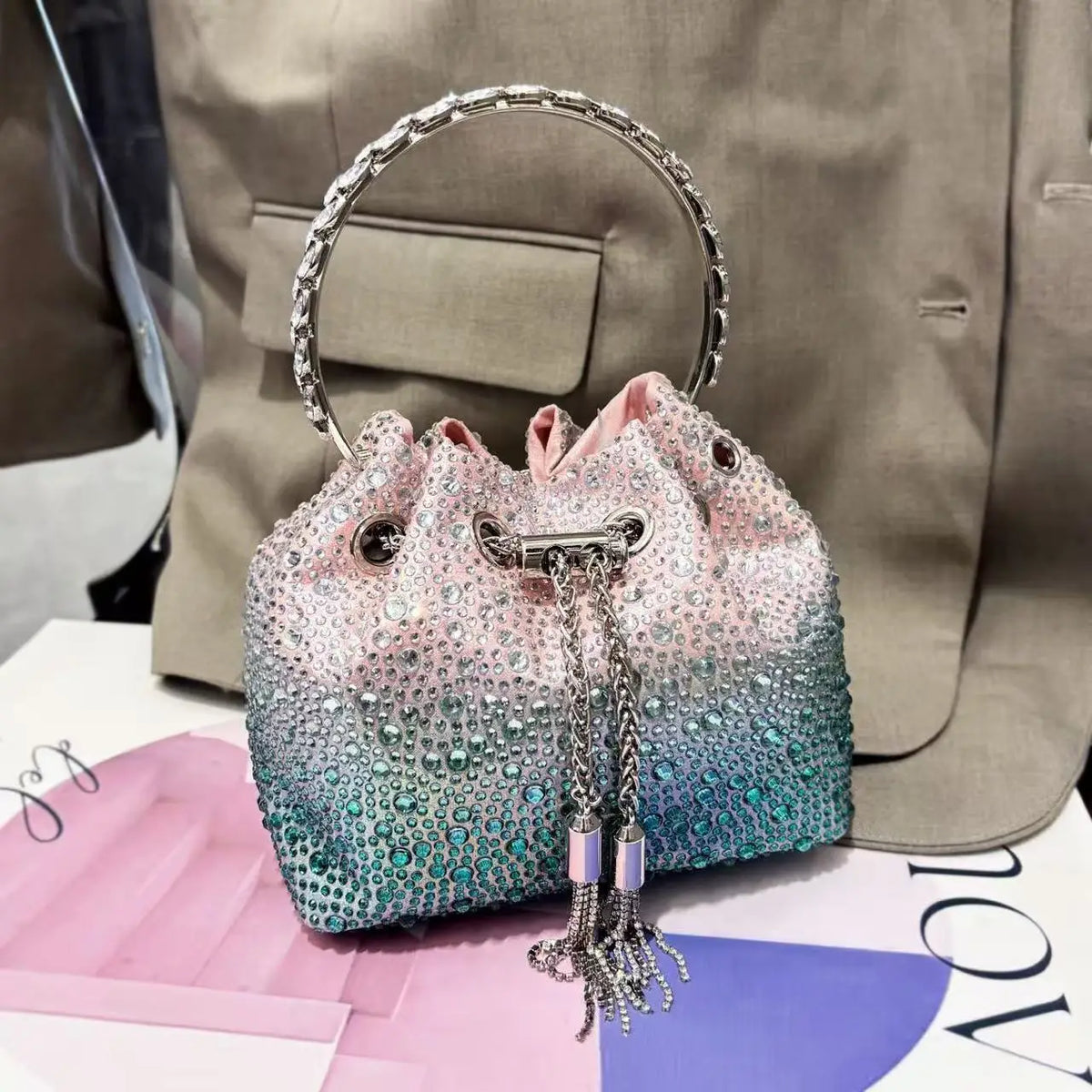 A pink and blue crystal-embellished evening clutch with a silver chain resting on a pink surface.