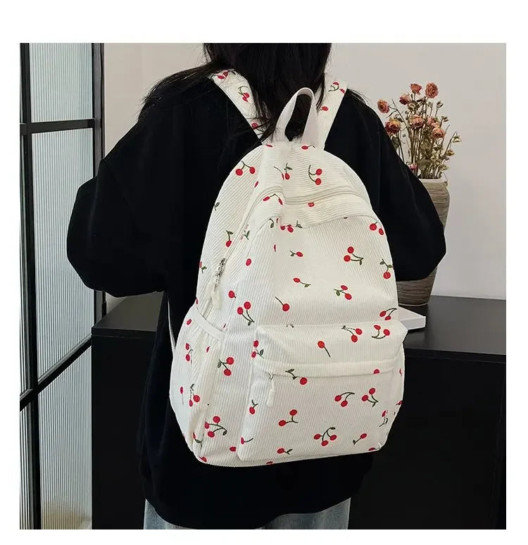 A white corduroy fashion backpack with a pattern of red cherries is worn by a person.