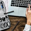 A hand is opening a gray travel cable organizer pouch filled with various electronic accessories next to an open laptop.