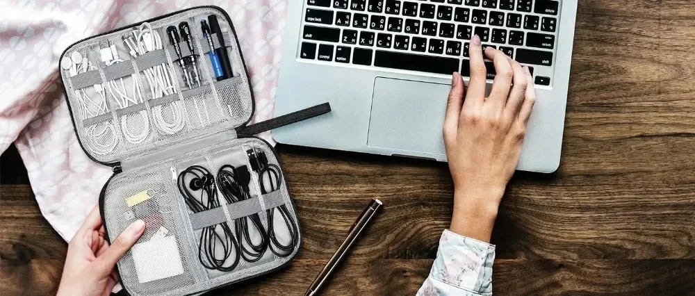 A hand is opening a gray travel cable organizer pouch filled with various electronic accessories next to an open laptop.