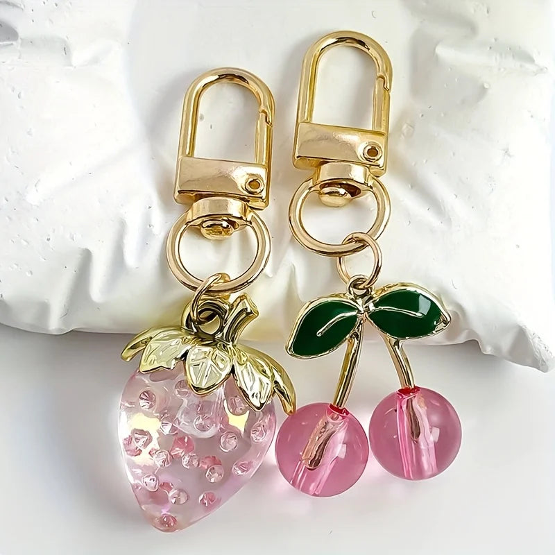 A set of gold-toned keychain charms featuring a transparent pink strawberry and cherry bag design.