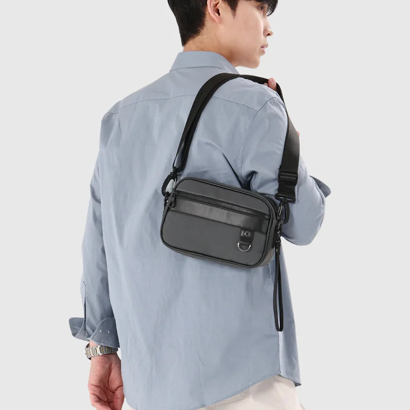 Una persona vistiendo una camisa azul claro lleva un bolso cruz de cuero PU negro con correa ajustable.