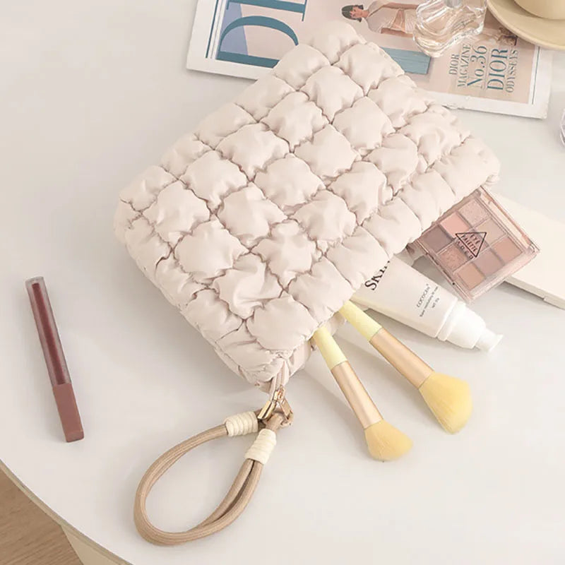 A quilted beige puffer makeup bag rests on a white surface next to makeup brushes and cosmetic products.