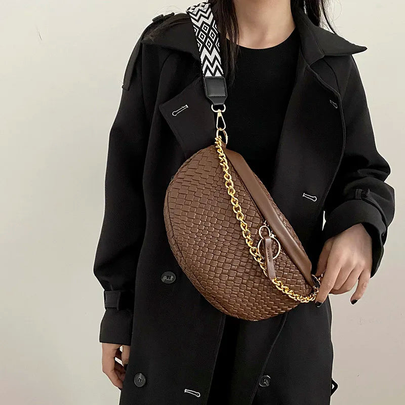 A woman holds a brown woven pattern waist bag with a gold chain strap under a black coat.