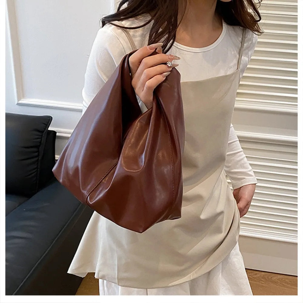 A woman holds a brown leather minimalist crossbody tote bag while wearing a cream-colored top.