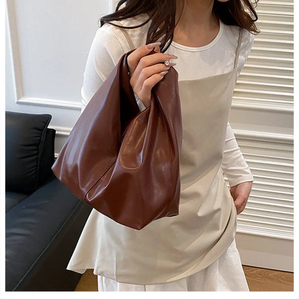 A woman holds a brown leather minimalist crossbody tote bag while wearing a cream-colored top.