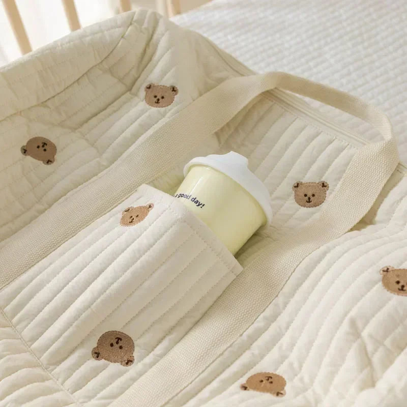 A beige quilted diaper bag tote with bear embroidery is shown resting on a white blanket.