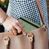 A hand is inserting an RFID blocking leather travel wallet into a tan leather handbag.