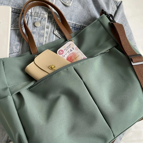 A light sage green nylon tote bag with brown leather straps and a flap pocket featuring a small tag.