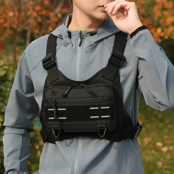 Eine Person trägt eine graue Kapuzenjacke und hält einen schwarzen taktischen Brustgurt-Rucksack für Outdoor-Ausrüstung hoch.