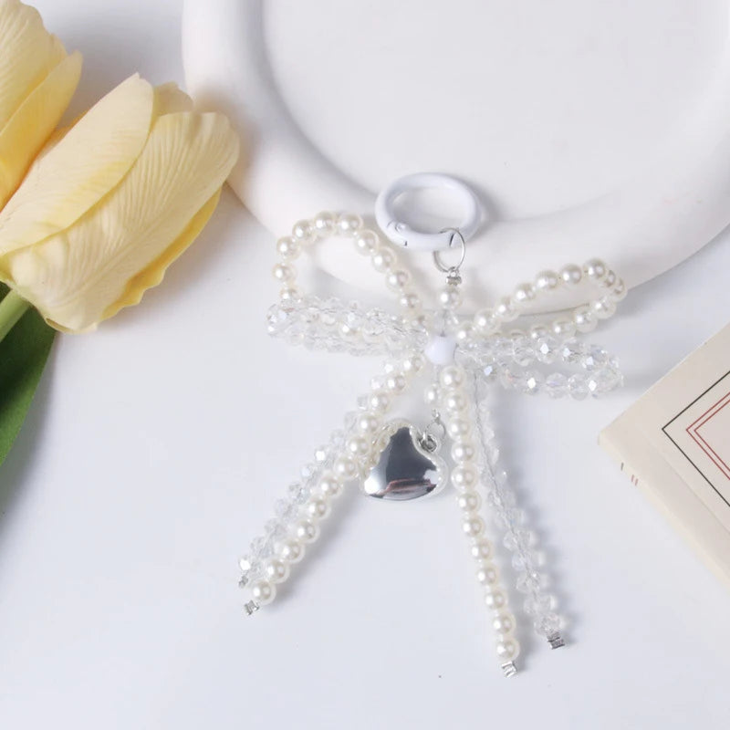 A clear pearl keychain with a bow design and a silver heart charm resting on a white surface next to a yellow tulip.