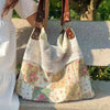 A light-colored patchwork shoulder bag with floral patterns is being held by a person outdoors.
