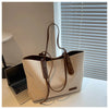 A beige canvas shoulder tote bag with brown leather straps resting on a white desk next to a laptop.
