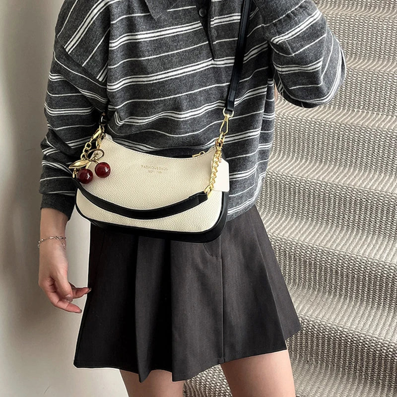 A person is holding a cream-colored shoulder bag with gold chain details and a red pendant against a striped background.