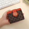 A hand holds an orange and brown monogrammed compact wallet with a leather flap.