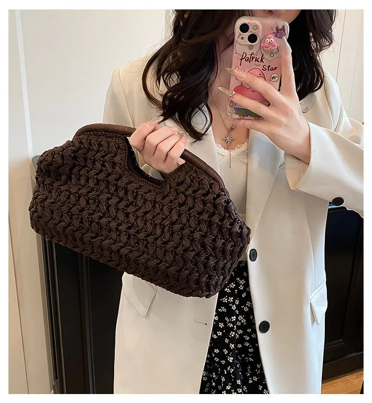 A woman holds a dark brown straw woven tote handbag against a white blazer and a patterned skirt.