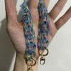 A hand holds a long, iridescent blue resin bag strap with gold-toned charms.