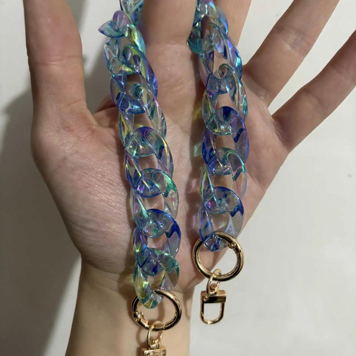 A hand holds a long, iridescent blue resin bag strap with gold-toned charms.