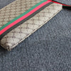 Beige gemusterte Clutch-Tasche mit roten und schwarzen Streifen liegt auf einer grauen Stoffoberfläche.