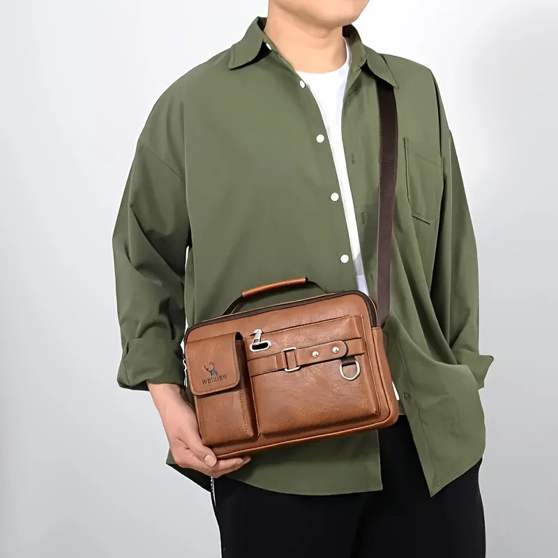 Un homme portant une chemise vert olive et un pantalon noir tient un sac bandoulière en faux cuir marron.