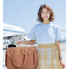 A young woman wearing a light blue shirt and plaid skirt stands next to a brown tote bag on an airport tarmac.