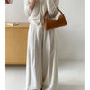 A woman wearing a cream-colored blouse and wide-leg trousers holds a brown faux suede crossbody bag.