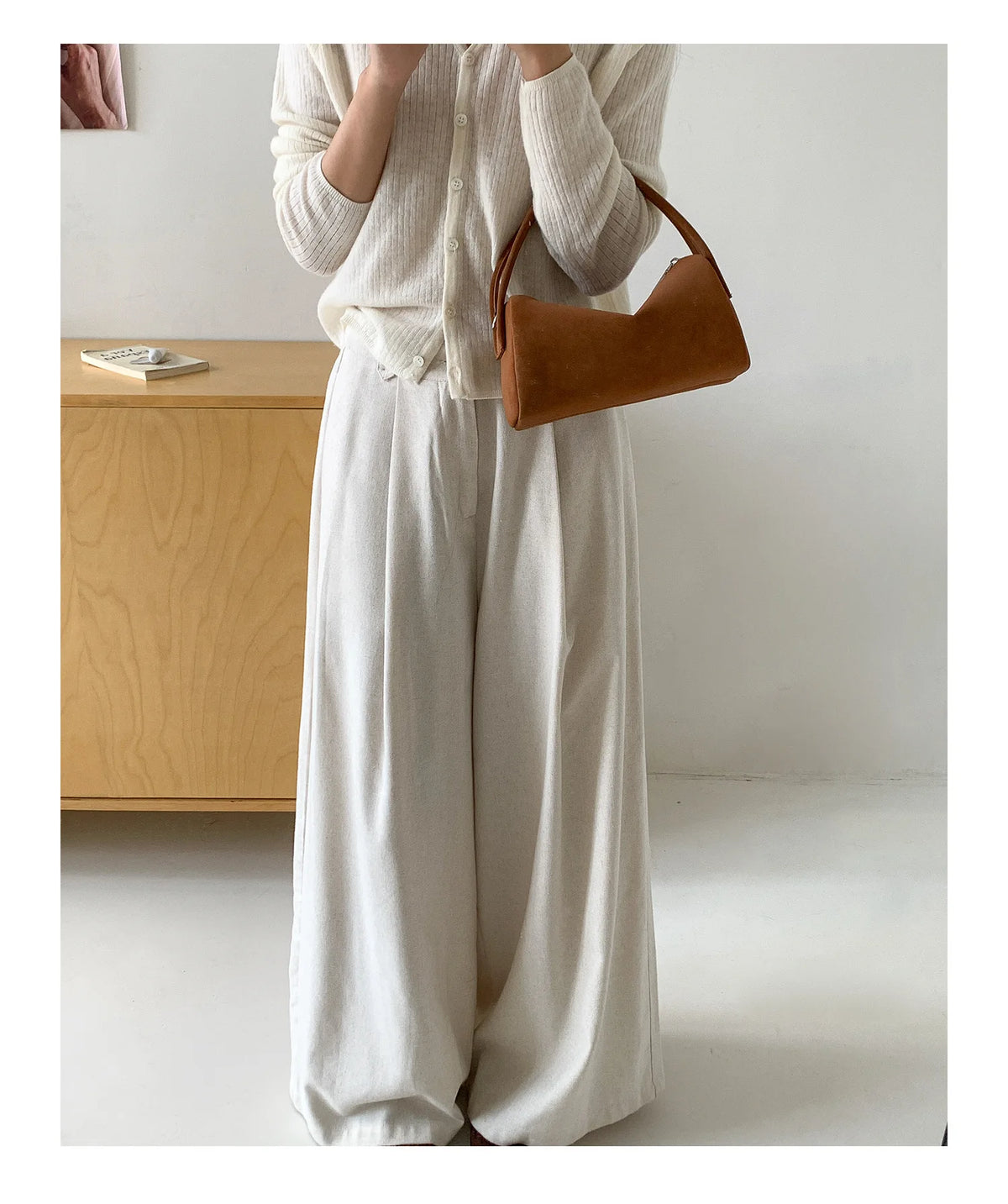 A woman wearing a cream-colored blouse and wide-leg trousers holds a brown faux suede crossbody bag.