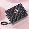 A black patterned letter zipper wristlet wallet with a gold logo resting on a light pink surface.