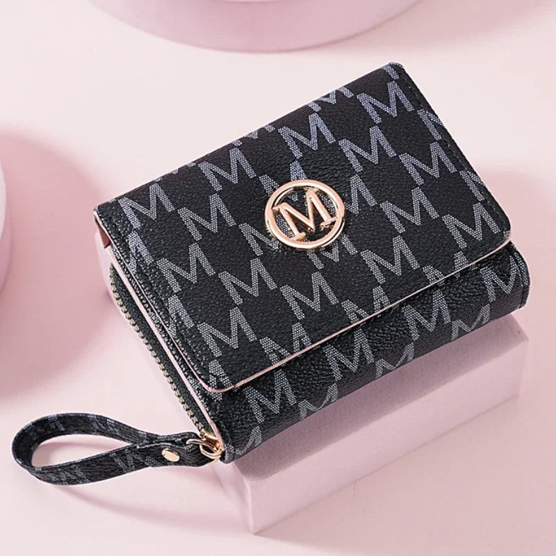A black patterned letter zipper wristlet wallet with a gold logo resting on a light pink surface.