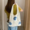 A woman is holding a white knitted tote bag with blue swirl and star embroidery against a light background.