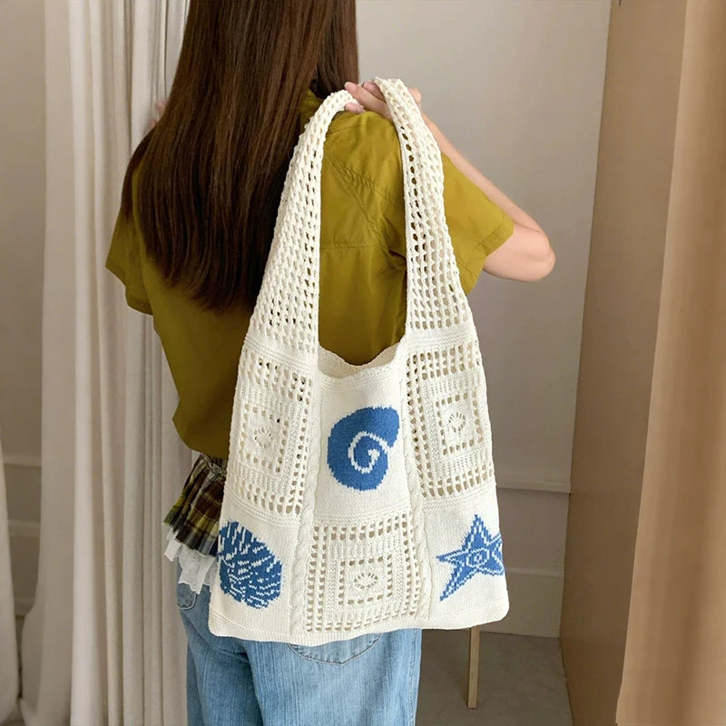A woman is holding a white knitted tote bag with blue swirl and star embroidery against a light background.