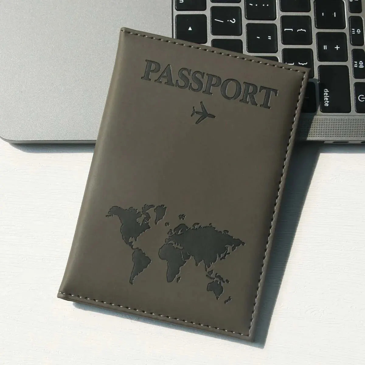 A brown leather passport holder with a world map design rests on a white surface next to a laptop keyboard.
