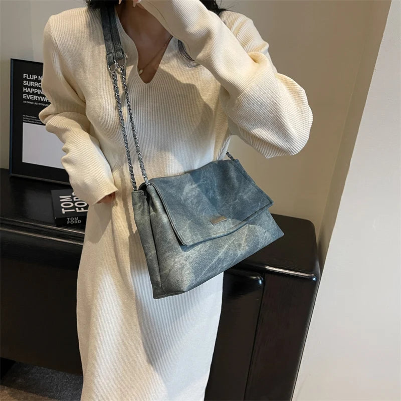 Una mujer modela un bolso de cuero gris texturizado con correa de cadena y un vestido largo de punto blanco.