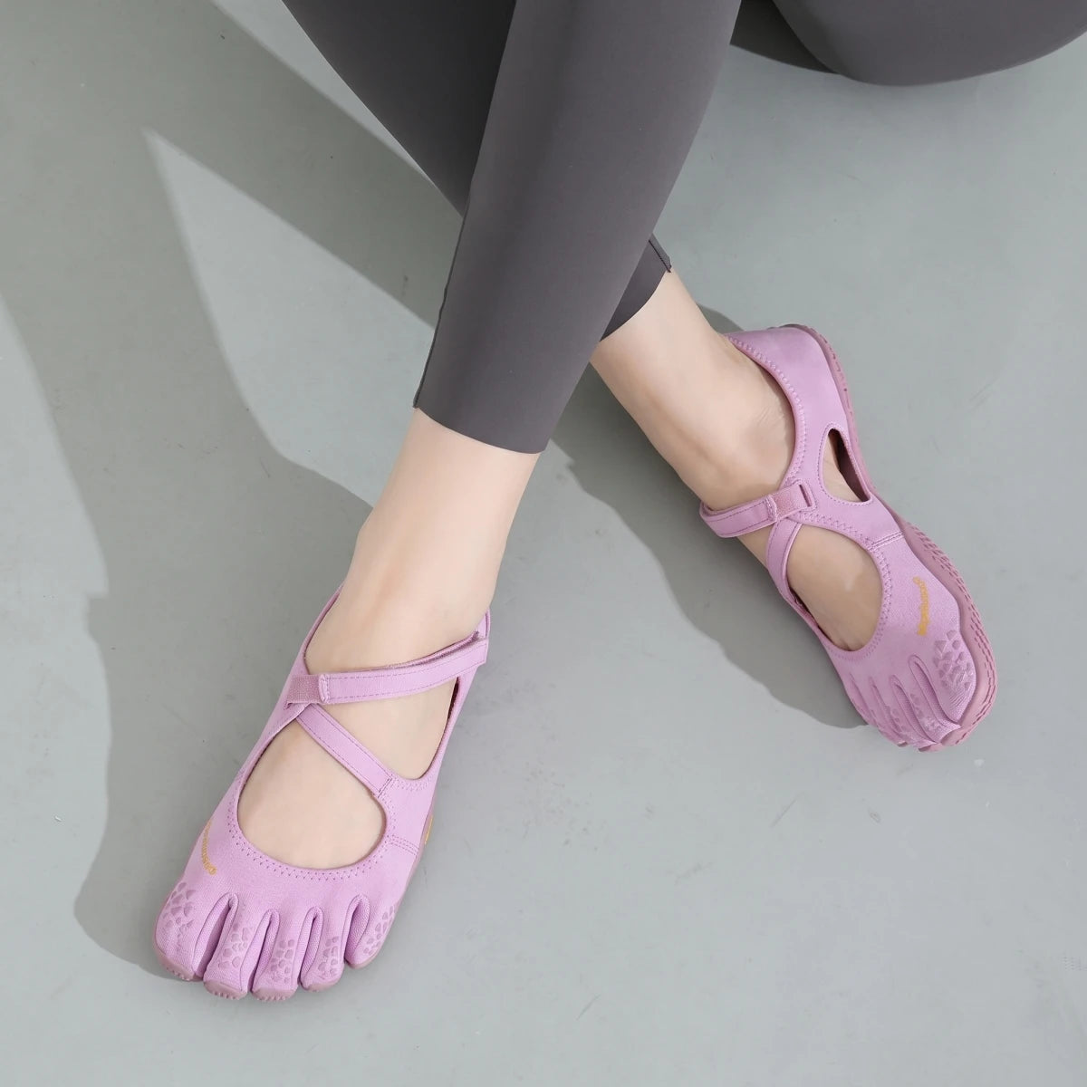 A close-up of a woman's foot wearing pink barefoot five-toe shoes with adjustable straps against a gray background.