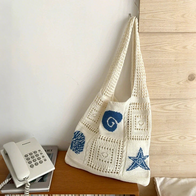A cream-colored knitted tote bag with blue swirl and star patterns is displayed next to a white calculator on a wooden surfac