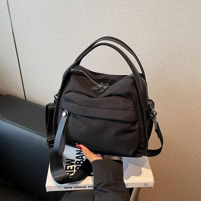 A black multi-pocket nylon crossbody bag with a top handle is displayed on a white surface.
