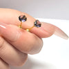 A hand holds a set of stainless steel ear stud earrings featuring purple and pink gemstone-like accents.