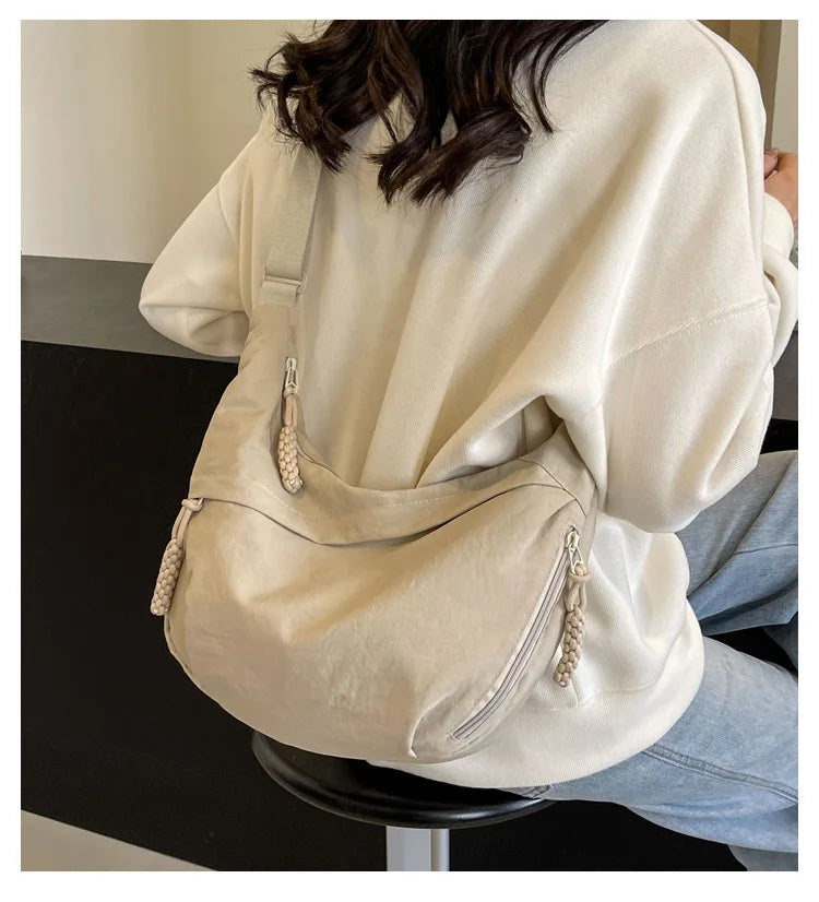 Eine Frau hält eine beige Nylon-Umhängetasche unter einem cremefarbenen Sweatshirt.