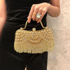 A hand wearing a ring holds an elegant gold diamond rhinestone evening clutch bag with golden tassels against a black dress.