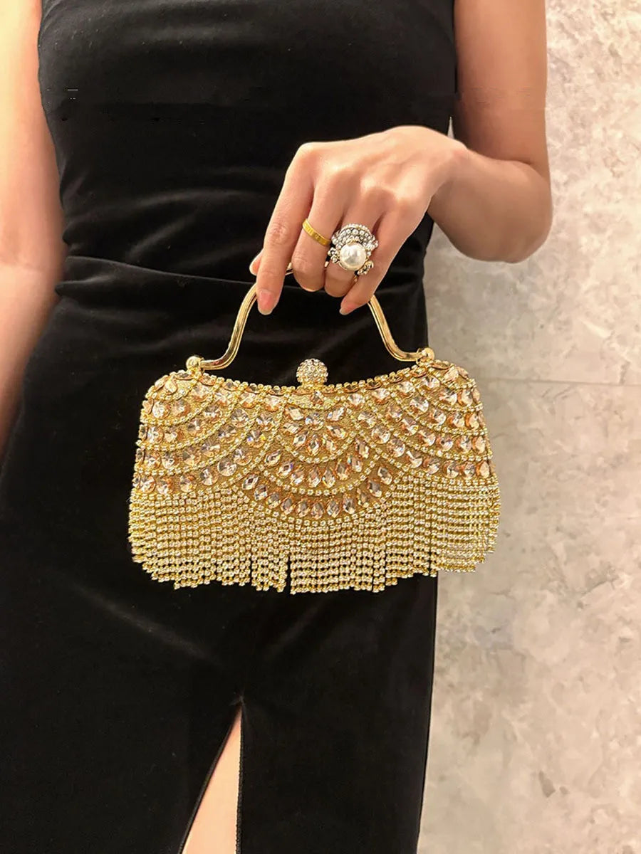 A hand wearing a ring holds an elegant gold diamond rhinestone evening clutch bag with golden tassels against a black dress.