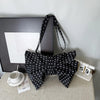 A black polka dot pleated bow crossbody bag displayed on a light surface next to a framed picture and a numbered card.