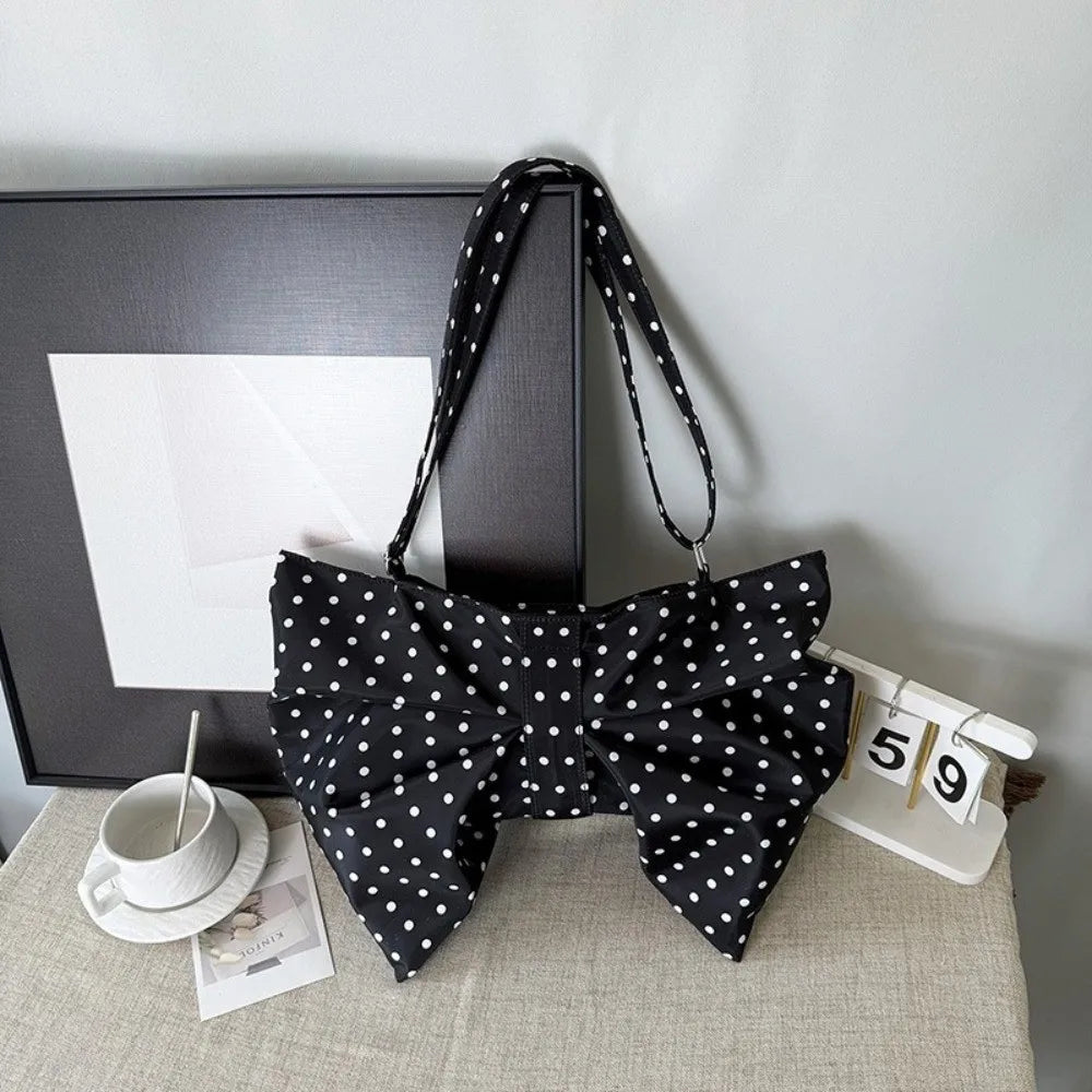 A black polka dot pleated bow crossbody bag displayed on a light surface next to a framed picture and a numbered card.