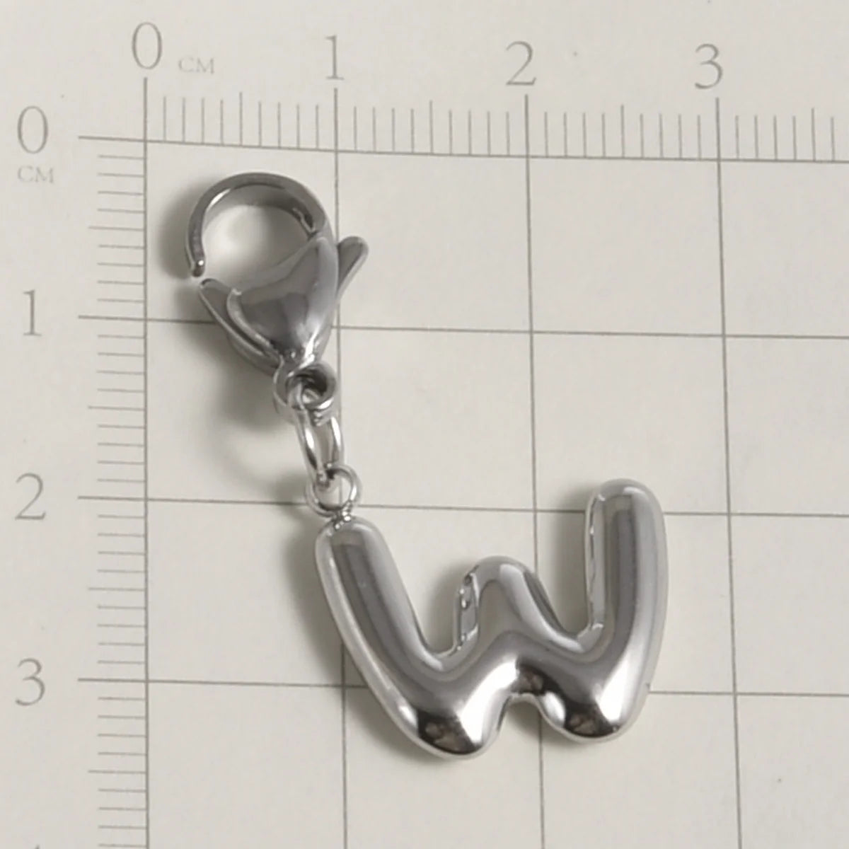 A silver stainless steel alphabet letter charm shaped like the letter W with a lobster clasp, measured against a ruler.