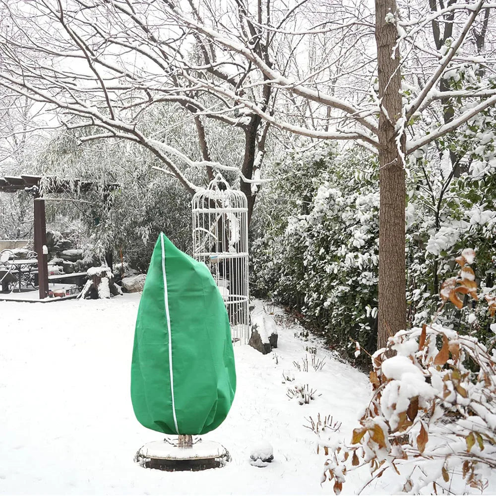 A green plant protection cover with a zipper and drawstring is displayed outdoors in a snowy garden setting.
