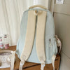 A light blue backpack with beige straps is displayed on a wooden surface next to a window.