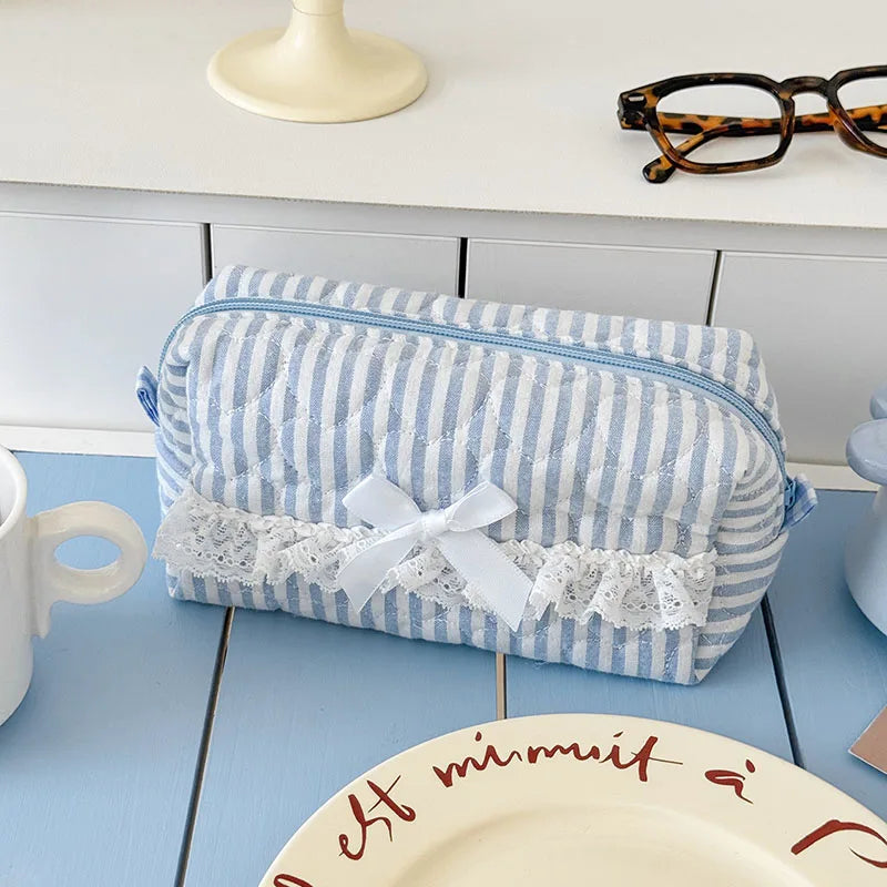 A striped blue and white cosmetic storage pouch with a white ribbon and lace trim resting on a light blue table.