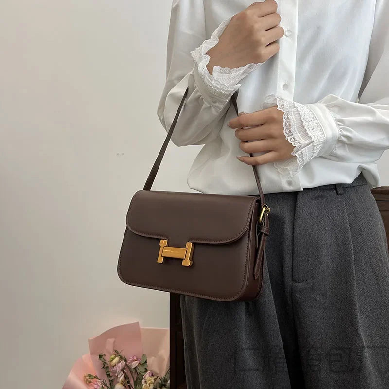 Braune Leder-Ein-Schultertasche mit goldener Schnalle, gehalten von einer Person in weißem Bluse und grauem Rock.